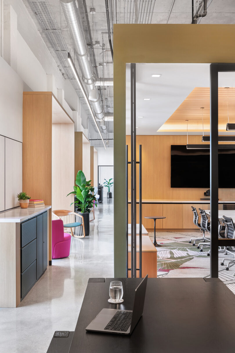 Modern office interior featuring a laptop on a foreground desk, with an open door leading to a conference room and colorful seating area. green plants add freshness to the space