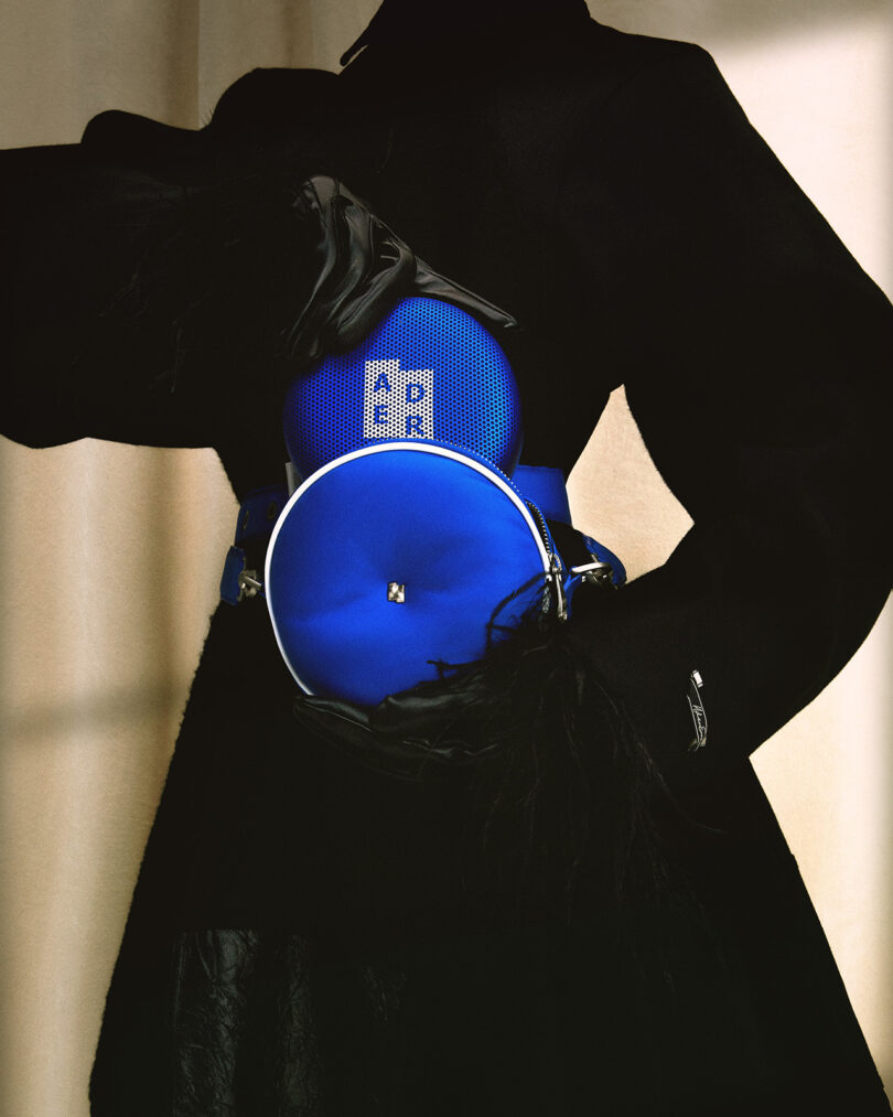 A person in a dark outfit holding Bang & Olufsen's Beosound A1 ADER ERROR Edition Bluetooth speaker, designed with a distinctive blue design.