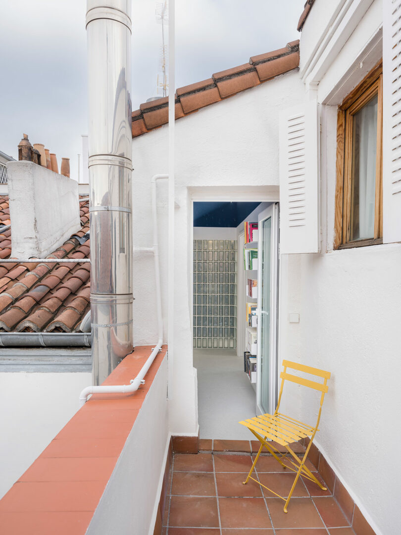 A small, open terrace with a single yellow chair, overlooking tiled rooftops, with an open door leading into a building.