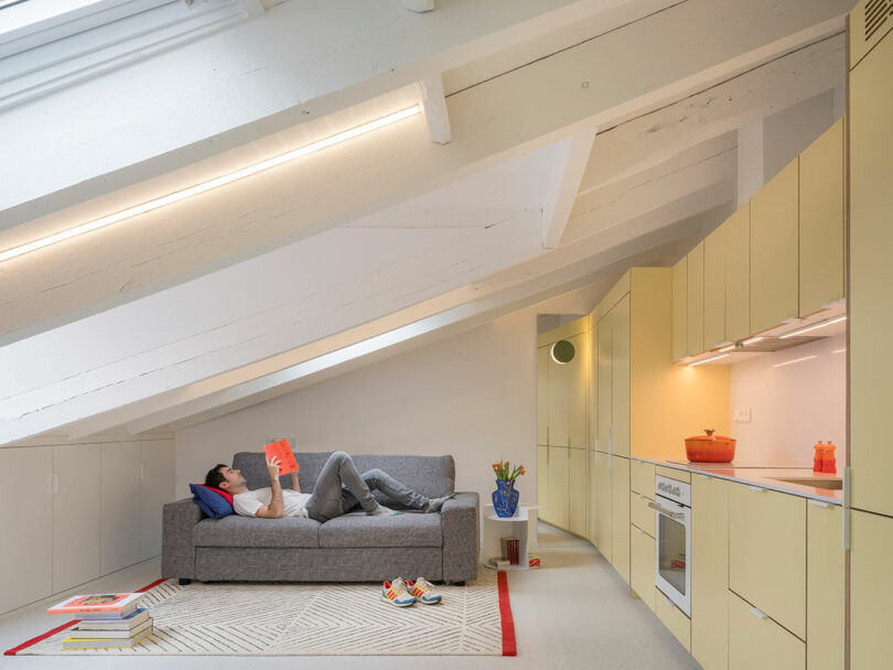 A person lounging on a gray sofa and reading a book in a modern loft with yellow cabinets and white beams.