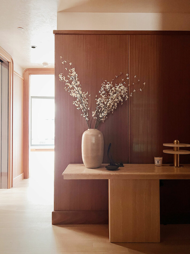 A warm-toned room in featuring a wooden table with a tall vase holding white flowers, beside a window and under soft lighting.