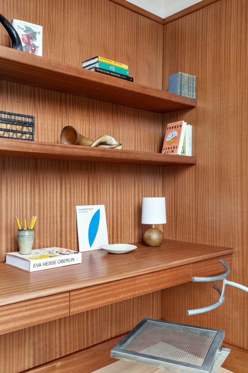 A cozy study corner in with wooden shelves holding books, decorative items, and a small white lamp, above a desk with stationery and a chair.