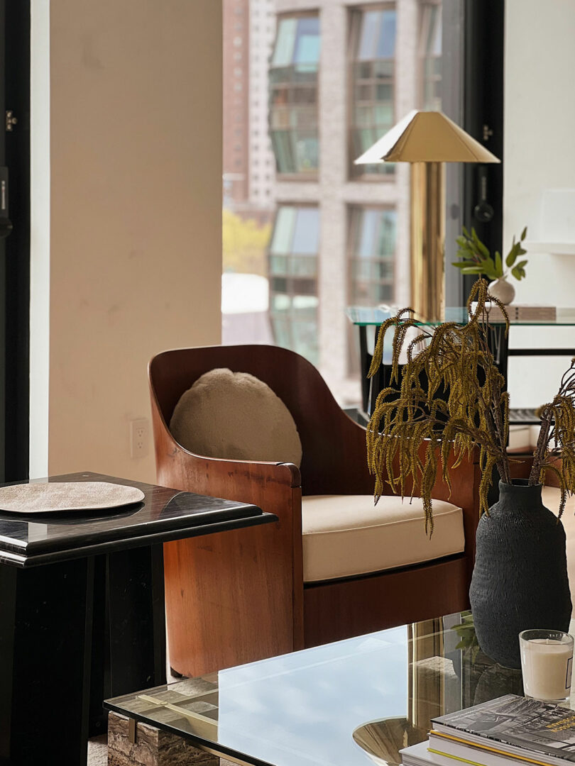 Modern living room corner in with a brown leather armchair, glass coffee table, and potted plants by a large window overlooking urban buildings.