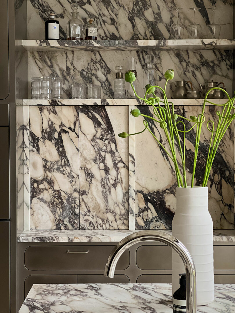 Modern kitchen with marble countertops and backsplash, featuring a white vase with green poppy stems and chrome faucet.