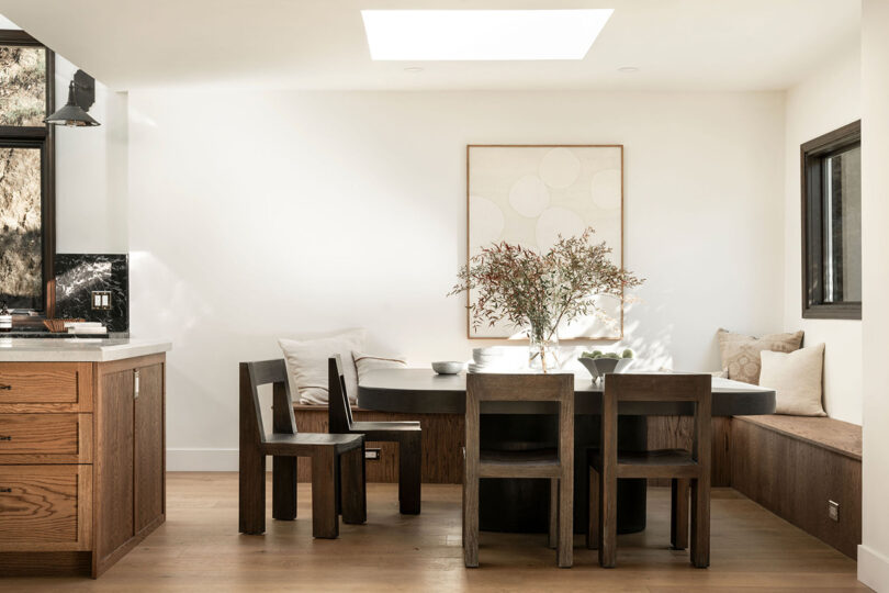 Modern dining room with wooden furniture, a plant on the table, and large windows showing outdoor trees.