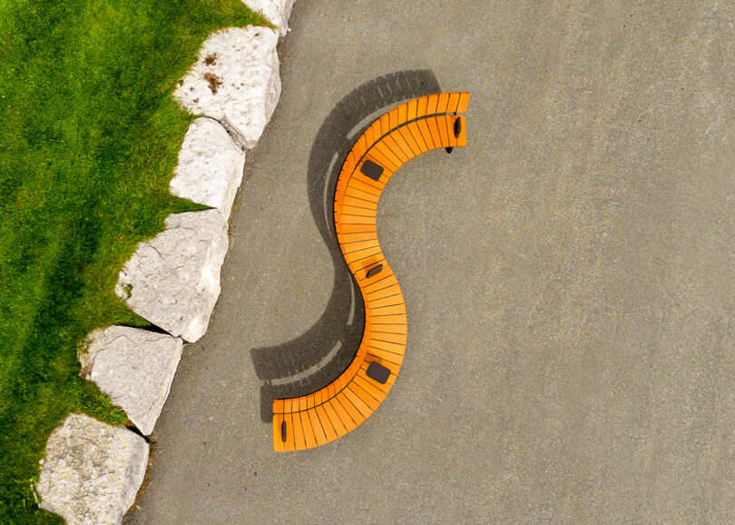 A curved wooden wooden bench with black metal supports, positioned on a concrete path beside a grassy area and stone wall.