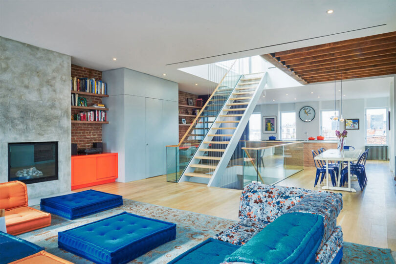 Modern living room with a mix of rustic and contemporary elements, featuring exposed brick, a central staircase, and vibrant furniture accents.