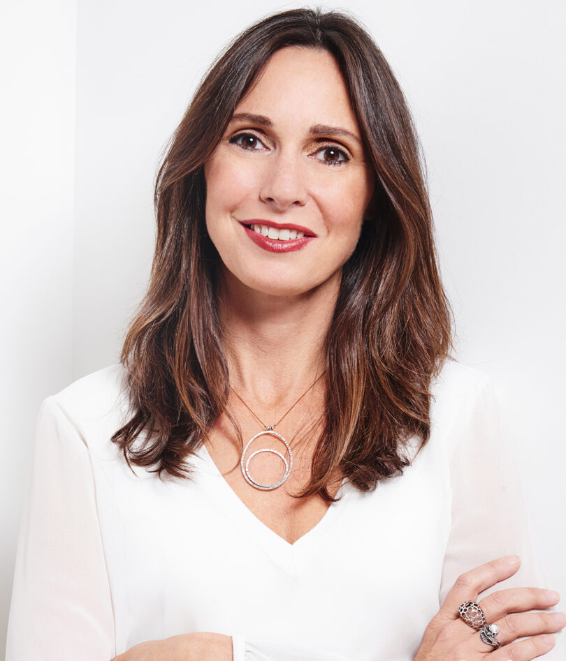 A professional portrait of a smiling woman with long hair, wearing a white blouse and a circular pendant necklace.