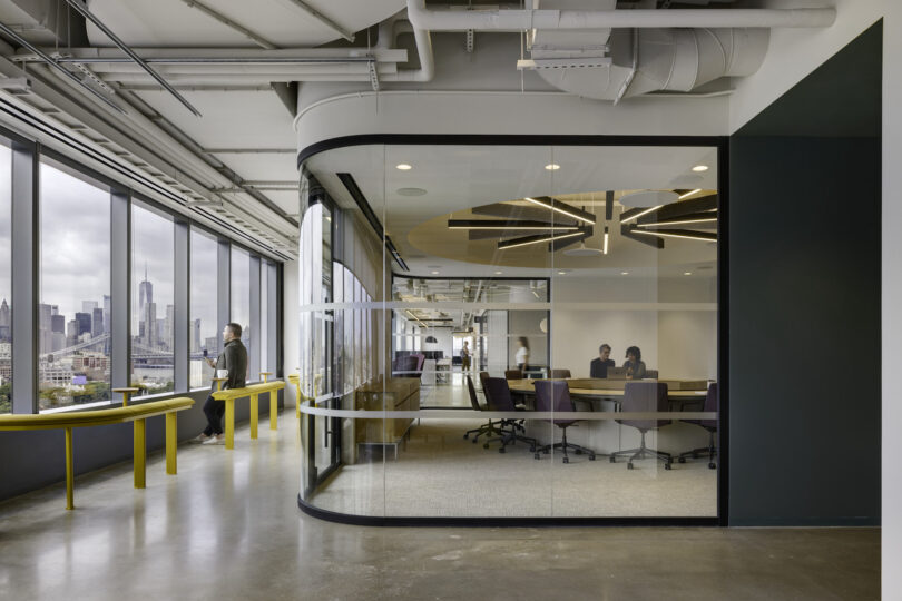 Modern office interior with glass walls, a man walking past a meeting room where several people are seated around a table, city skyline visible through large windows