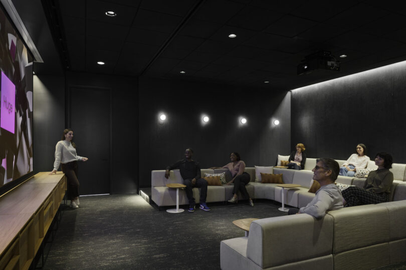 A woman presents on a screen in a modern, dimly-lit conference room with attentive listeners seated on sofas