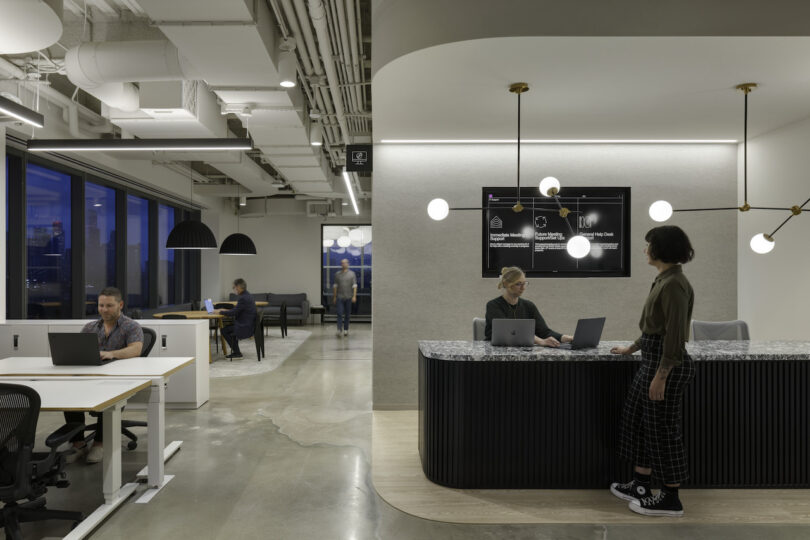 Modern office at dusk with employees working at desks and a reception area, city view through large windows, stylish interior with soft lighting