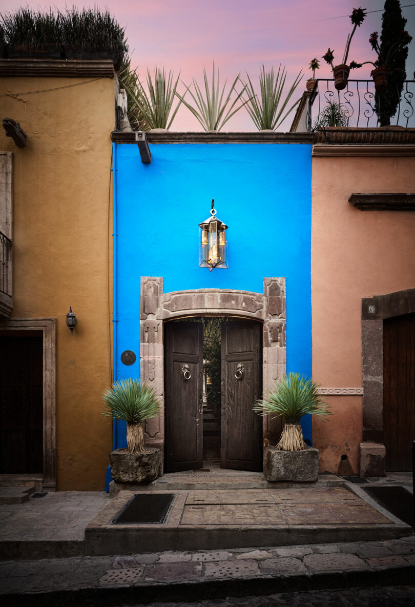 A vibrant blue wall with a hanging lantern above a wooden doorway, flanked by green potted plants and rustic stone steps, between two orange buildings.