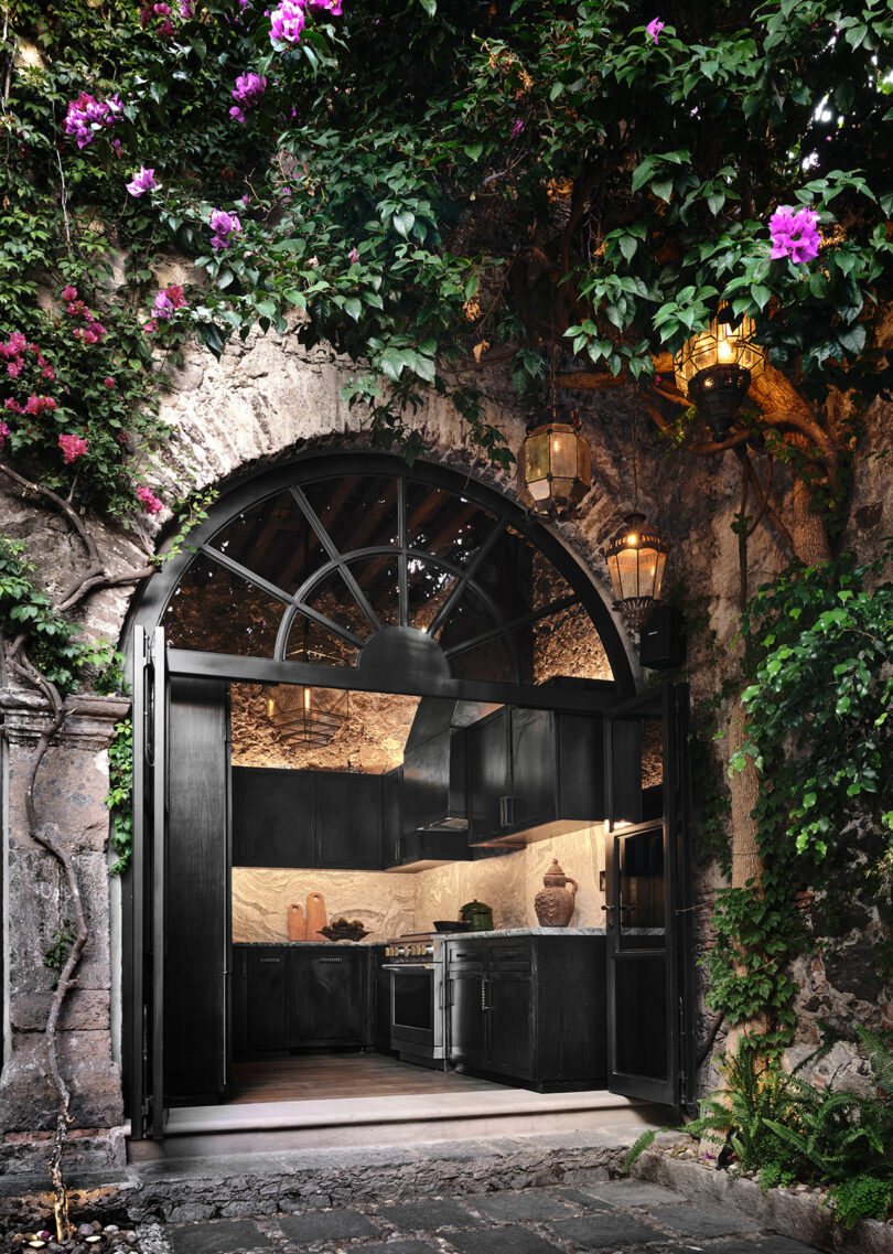 A kitchen built into a rustic stone archway, surrounded by lush green vines and pink flowers, with atmospheric lanterns hanging above.