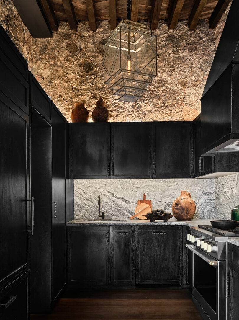 Modern kitchen with cabinets and marble countertops, featuring a rugged stone ceiling, wooden beams, and a geometric hanging light fixture.