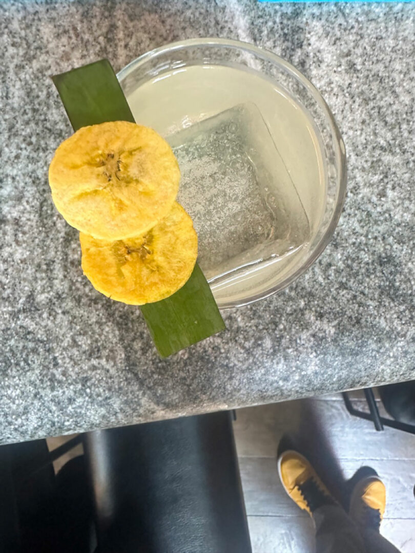 A glass cup on a kitchen counter with two banana slices garnishing the top, viewed from above with someone's feet visible in yellow shoes.