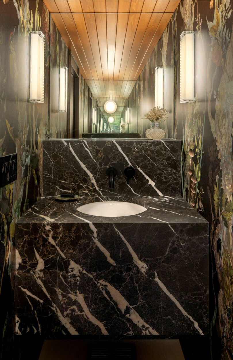 An elegant bathroom with a black marble vanity, white sink, and intricate floral wallpaper, complemented by warm lighting and vertical sconces.