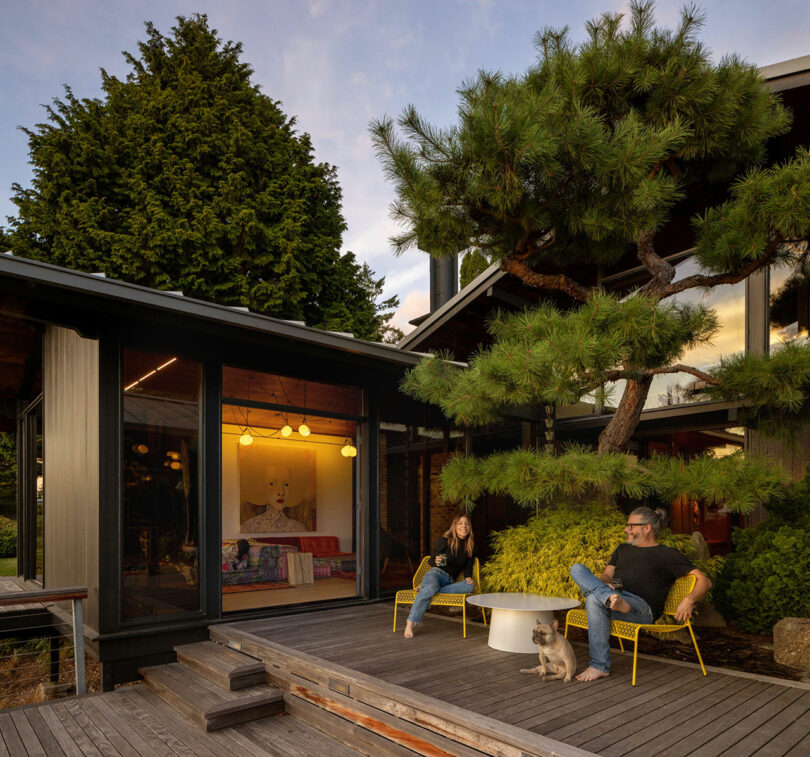 A couple relaxing on a wooden deck outside a modern house with large windows, surrounded by lush trees, with a small white dog near them.