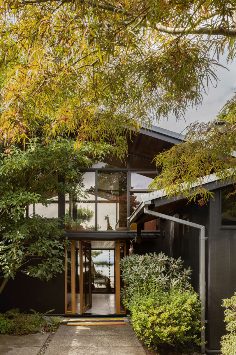 A modern home entrance with glass doors and wooden frames, surrounded by lush trees and plants, leading towards a body of water.