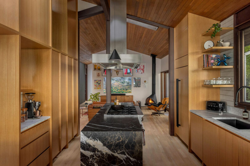 Modern kitchen with dark marble countertops and wooden cabinets, leading to a living area with a fireplace and mid-century modern furniture.