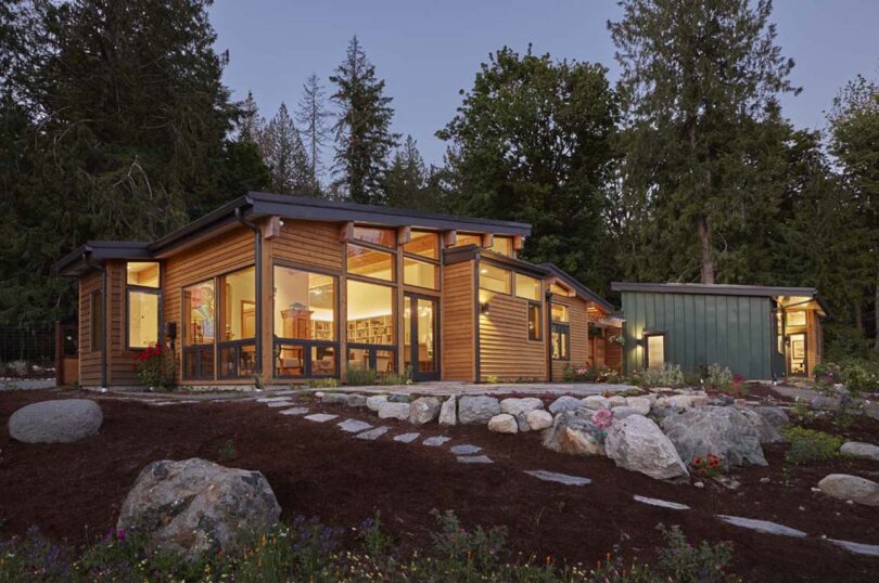 Modern house with large windows and wooden exterior illuminated at twilight, featuring a landscaped garden with a stone pathway.