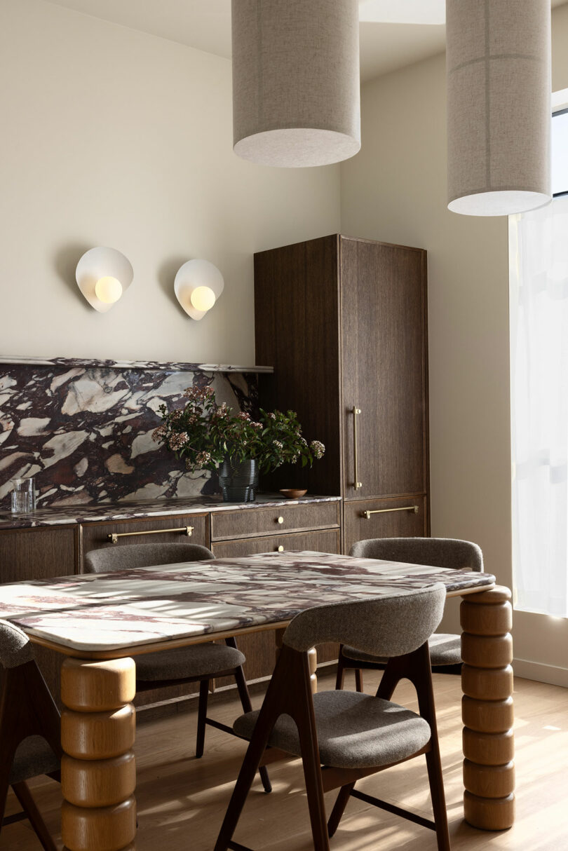 Modern dining room with a dark wood table, upholstered chairs, and unique cylindrical lighting fixtures, set against a light wall and marble detailing.