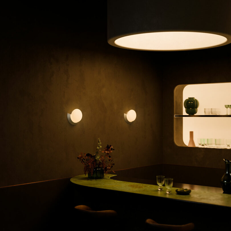 Dimly lit dining area with a round table under a large pendant light, two wall sconces, and a shelved alcove with decorative items.