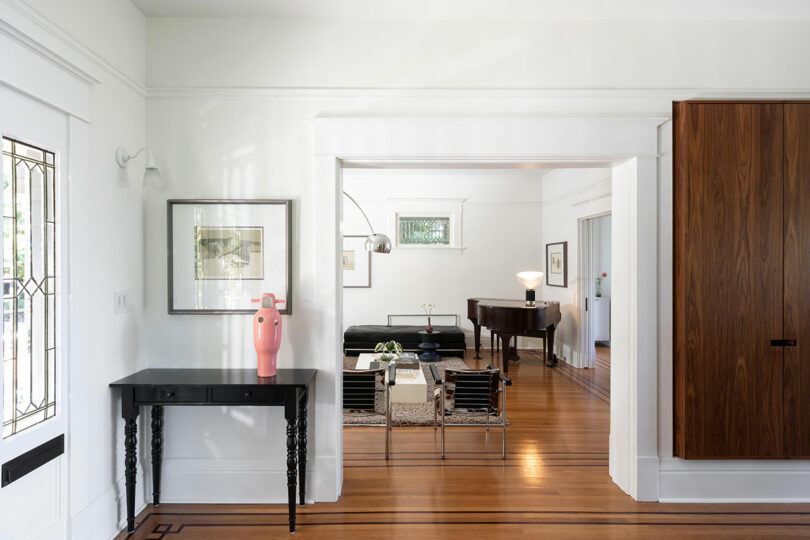 Bright living room with wooden floors; features a black console table, pink sculptural art, and an open wooden door leading to a room with a piano and black sofa.