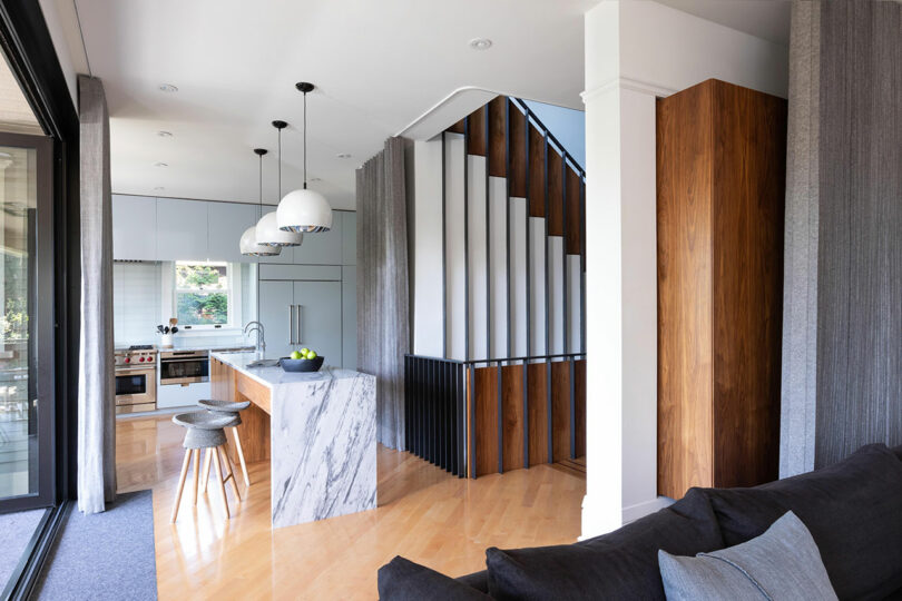 Modern kitchen with marble island adjacent to a living area with a gray sofa, and a staircase with wooden and white vertical paneling.