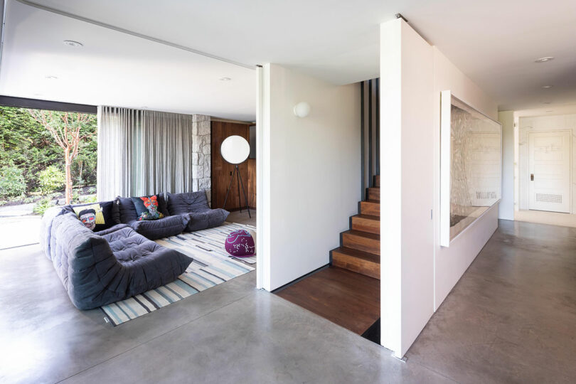 Modern spacious living room with large gray sectional sofa, stairs leading up behind a partial wall, and floor-to-ceiling windows showing outdoor greenery.