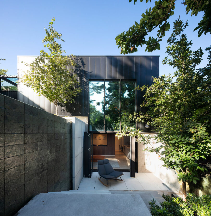 Modern two-story house featuring large glass windows, surrounded by lush greenery and a small outdoor seating area.