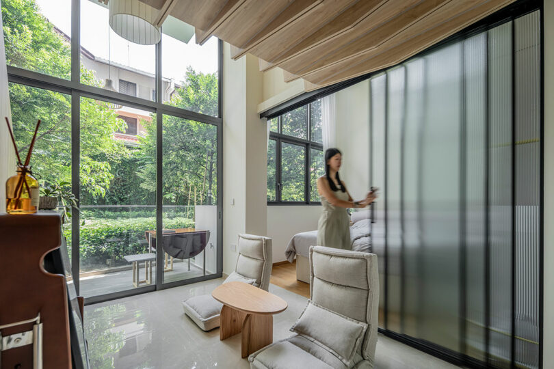 angled interior view of small modern apartment with two chairs on floor and woman pushing sliding glass doors open to bedroom