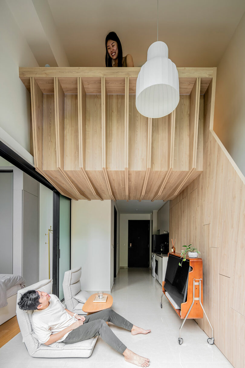 A woman in a raised wooden loft smiles down at a man lounging on a chair in a modern, wood-paneled room with minimalist decor.