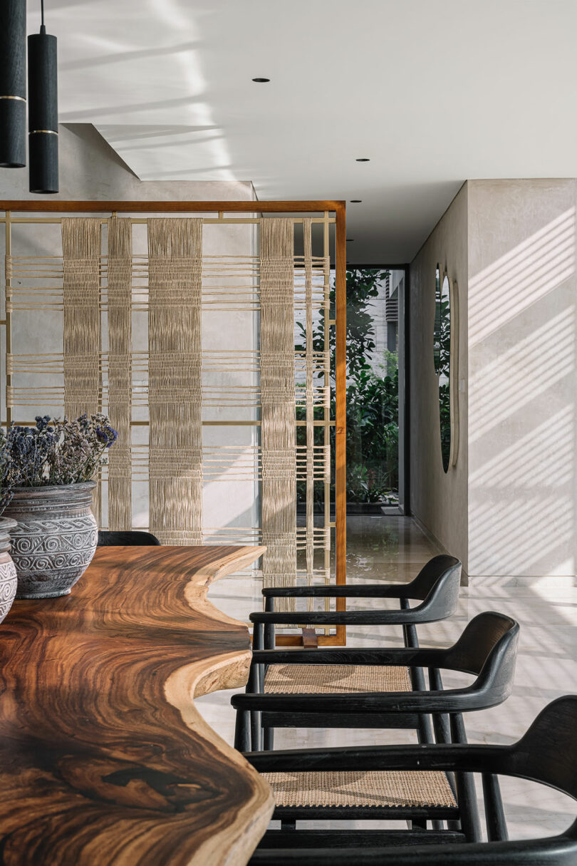 Modern interior featuring a wooden slab table, black chairs, woven room dividers, and a view of a lush garden through floor-to-ceiling windows.