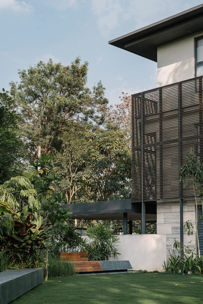 Modern house exterior with lush garden, featuring wooden slat details and large windows, surrounded by tropical plants and a small lawn area.