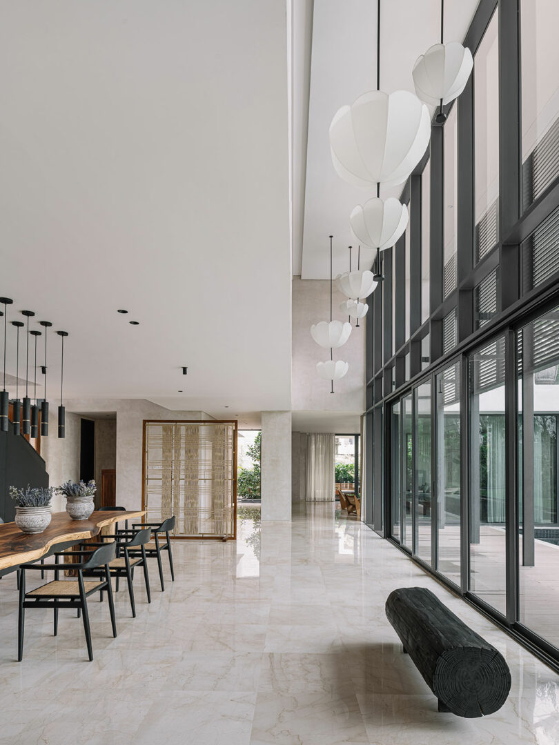 Modern airy lobby with high ceilings, marble floors, large windows, unique balloon-like hanging lights, a wooden dining table, and a cylindrical log bench.