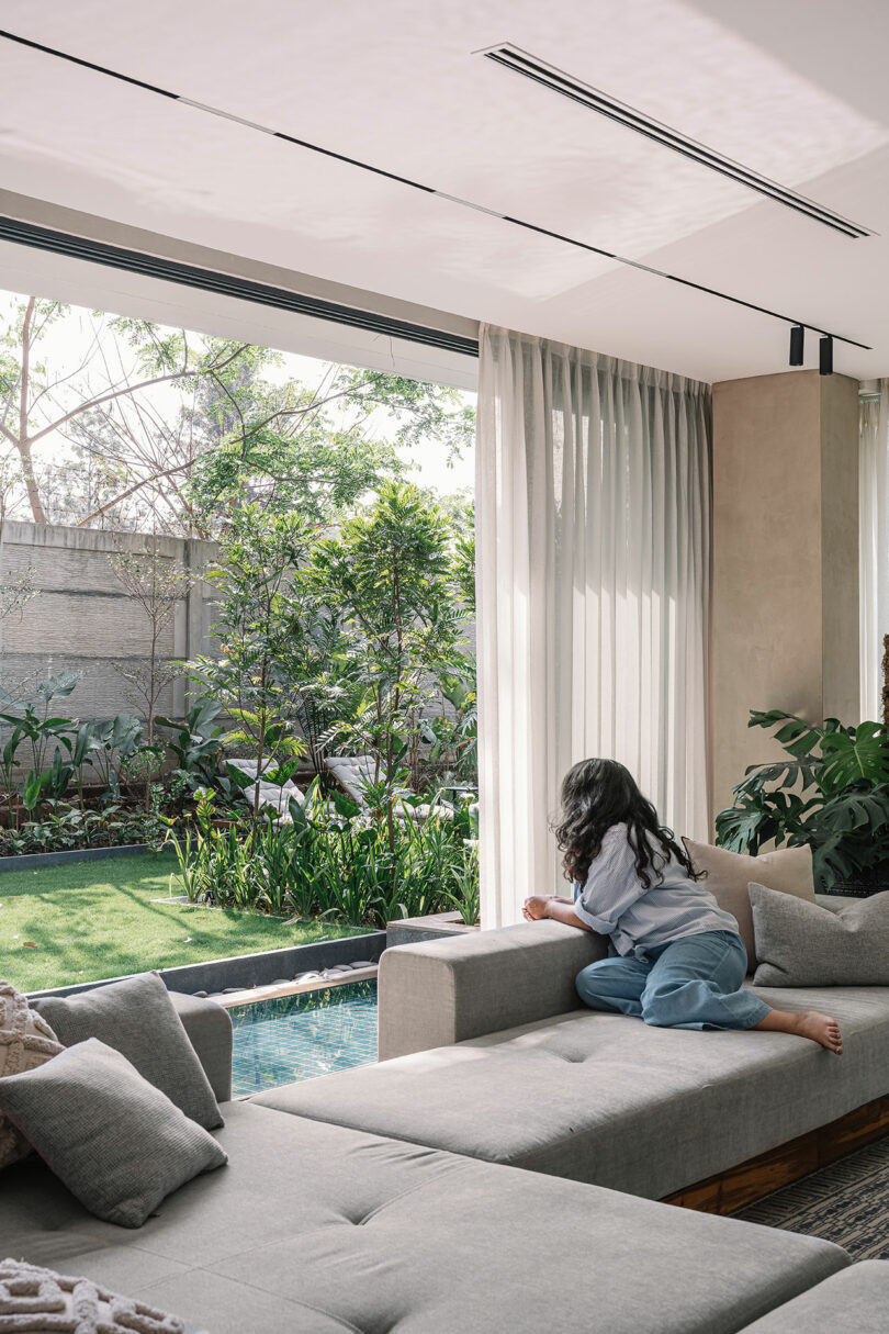 A woman sits on a couch reading a book, facing large windows that overlook a serene garden with a pond.